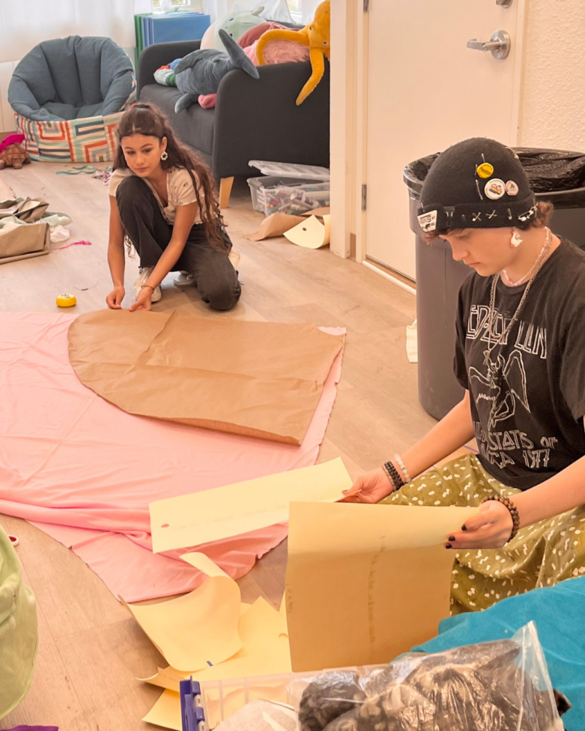 Two campers work on sewing projects indoors, using paper patterns and fabric spread out on the floor, surrounded by plush toys and cozy furniture.