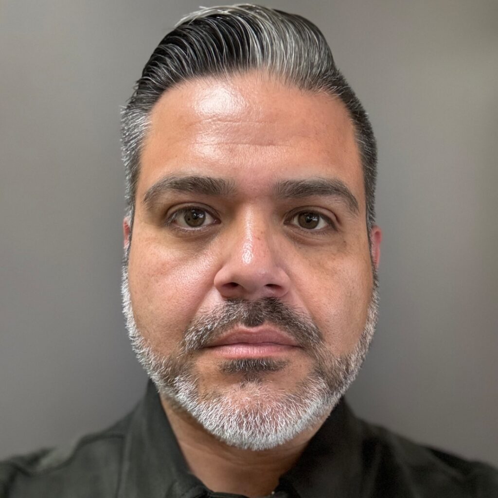 a headshot of a man with brown eyes and salt and pepper grey hair slicked over with a grey shortly clipped beard.