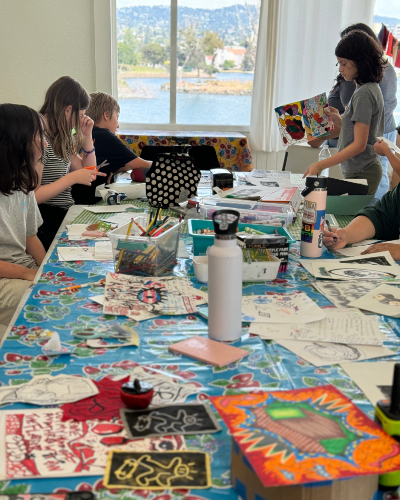 A table filled with various art supplies, including paints, brushes, markers, and sketchbooks, ready for creative projects. Students working busily in the background.