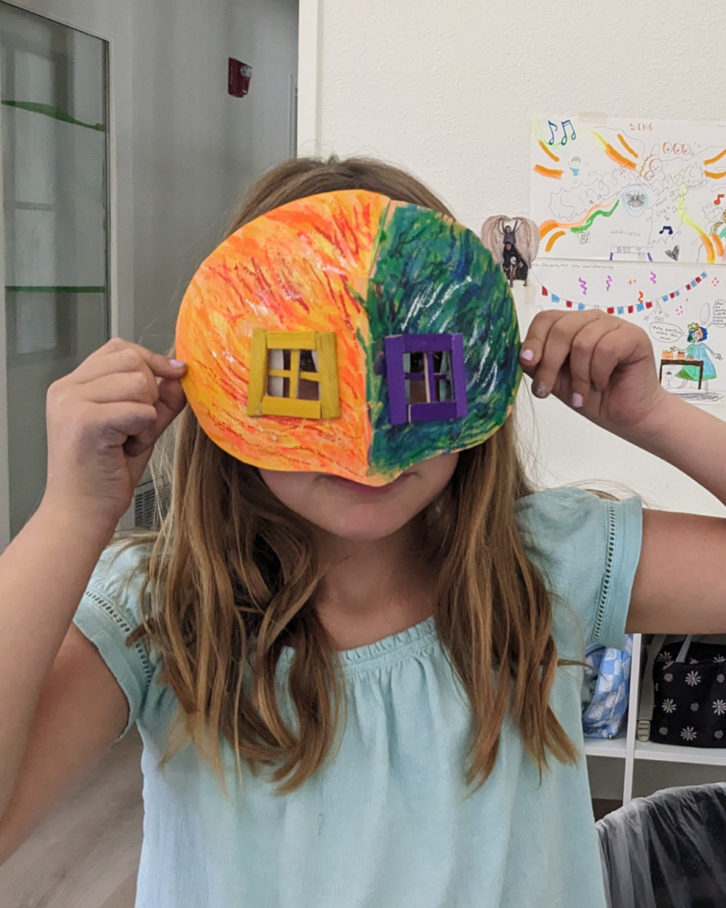 Camper holds up a handmade half orange, half green mask with window panes for eyes for her face.