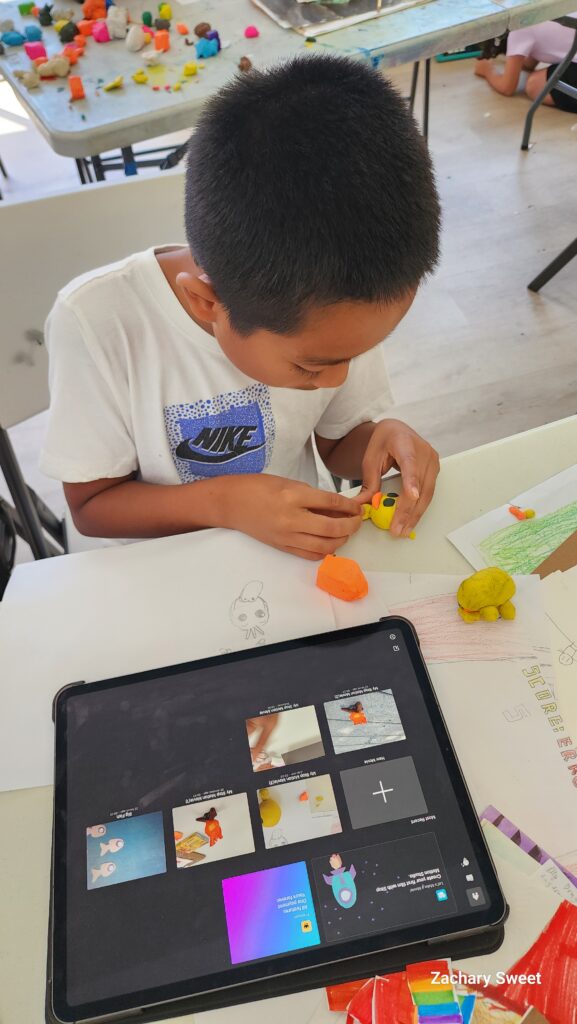 A camper is making a small duck head our of yellow clay for his stop motion movie. An iPad with a video editing app open is in the foreground.
