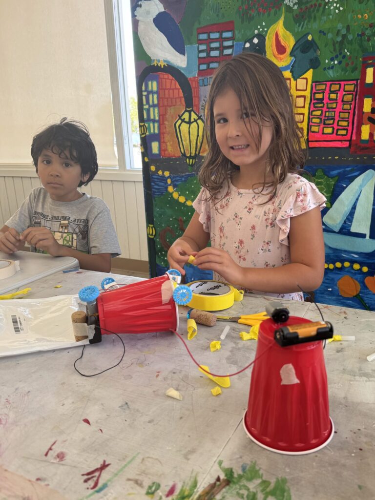 A smiling camper is taping batteries and wires to a red plastic cup to make a scribble bot: a small battery operated robot with markers for legs that can self-propel and doodle on paper.