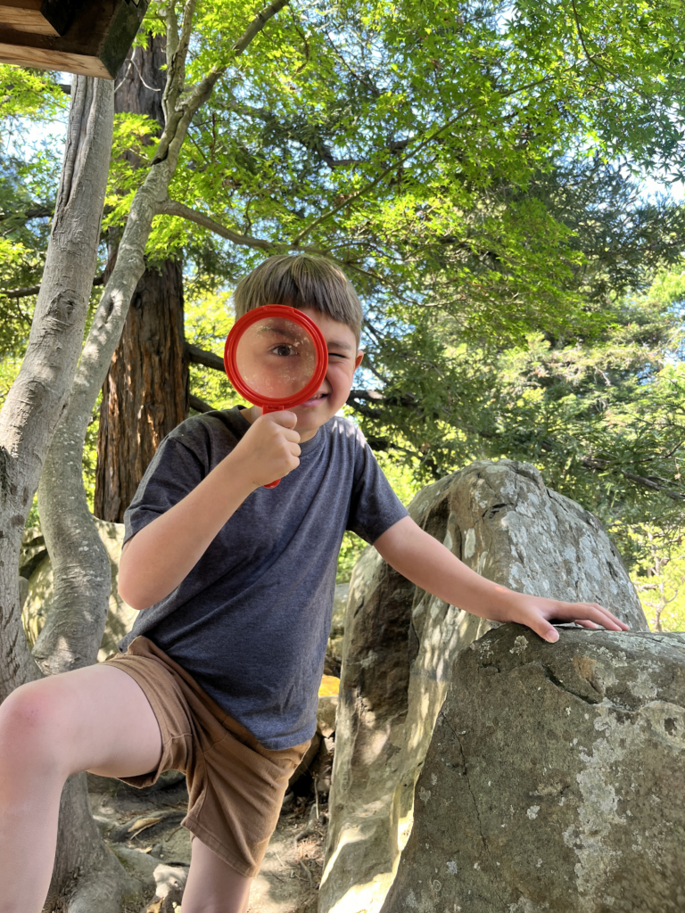 A boy is posing with a magnifying glass over his eye in the Lake Merritt gardens, surrounded my greenery, standing on a big rock.