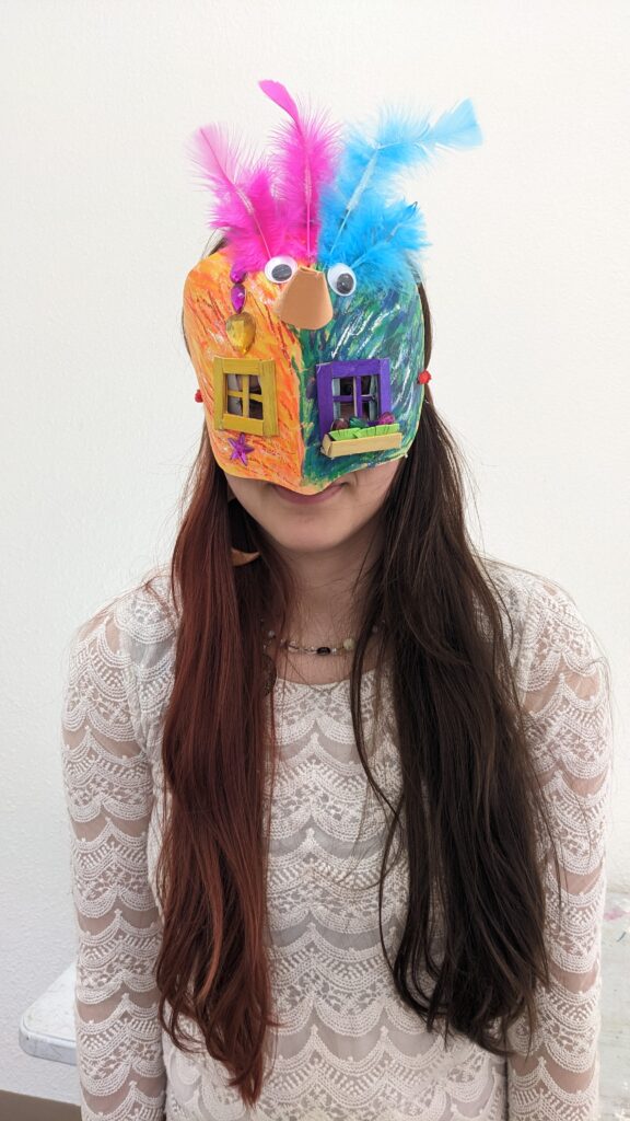 A camper is wearing a mask. The mask is half orange, half green mask with window panes for eyes. On the forehead area there are half pink and half blue feathers, a pair of googly eyes, and a beak made from a cone of orange felt.