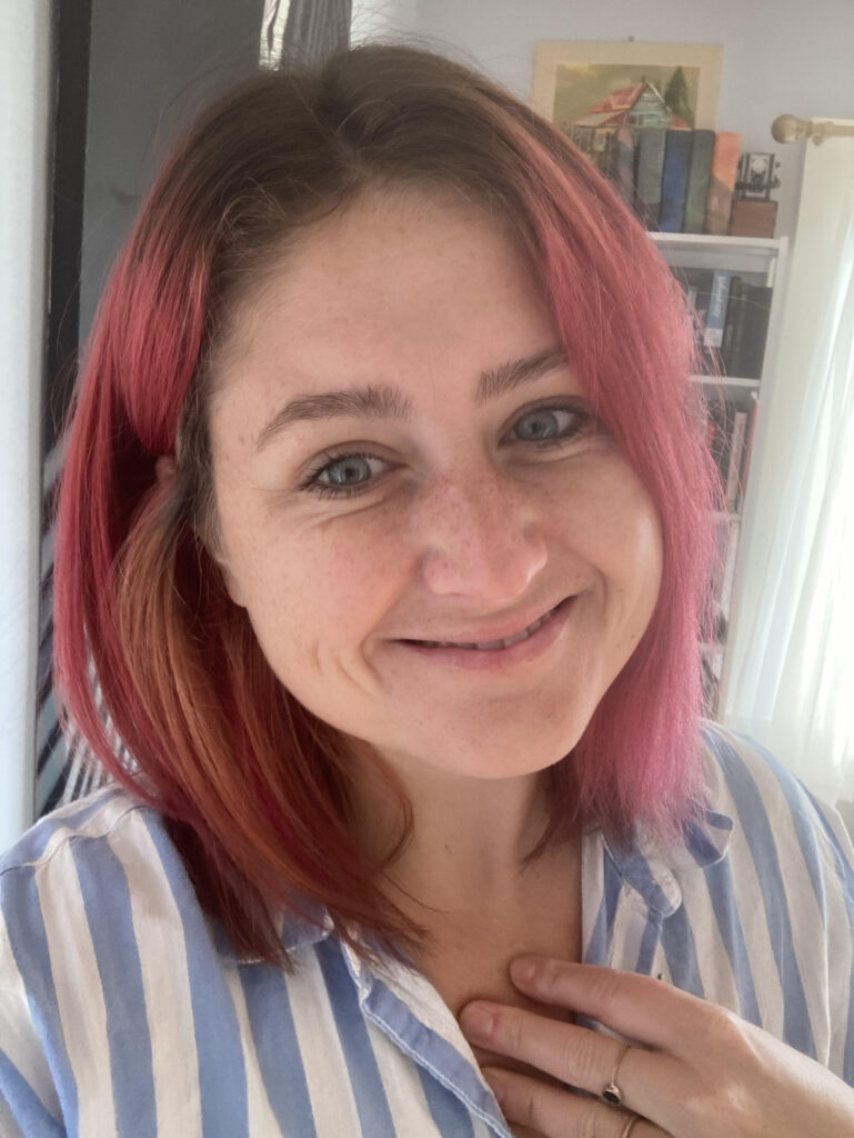Headshot of a white woman with blue eyes and pink hair, smiling. 