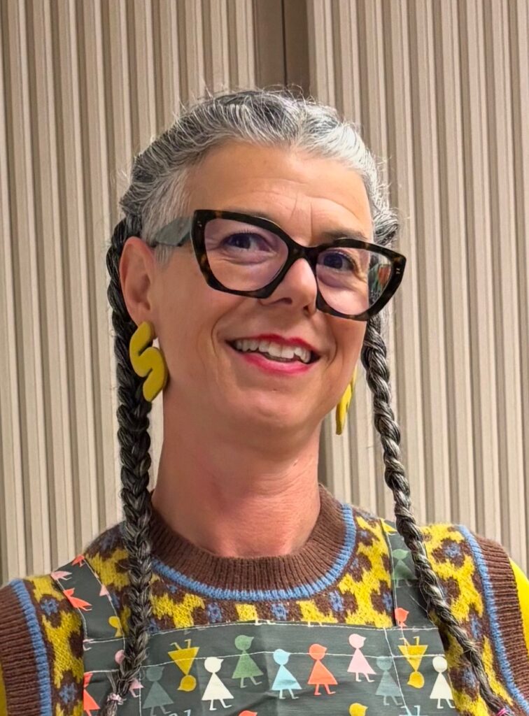 A smiling photo of a white woman with grey hair in braids and glasses with chunky yellow earrings on and a colorful top.