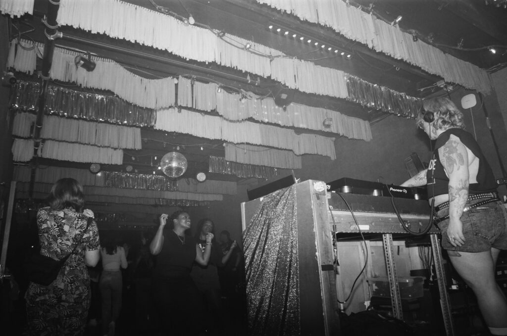 Black and white photo of people socializing and dancing in a lively club atmosphere. A tattooed DJ is in the foreground. Streamers and a disco ball hang from the ceiling in the background.